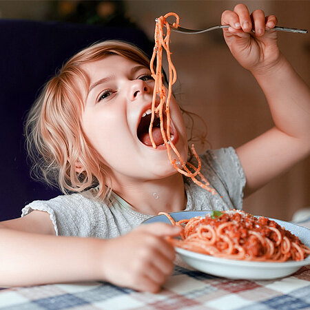 Ein Kind in einem grauen T-Shirt isst Spaghetti Bolognese. Es hält einige Spaghetti mit der Kabel in die Luft und versucht, diese mit dem geöffneten Mund zu schnappen.